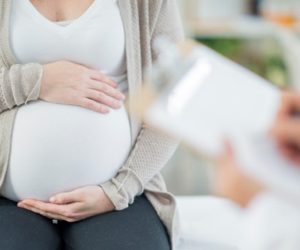 A pregnant Caucasian woman is indoors in a doctor's office. Her female doctor is wearing medical clothing. The woman is holding her stomach which the doctor writes on a clipboard.