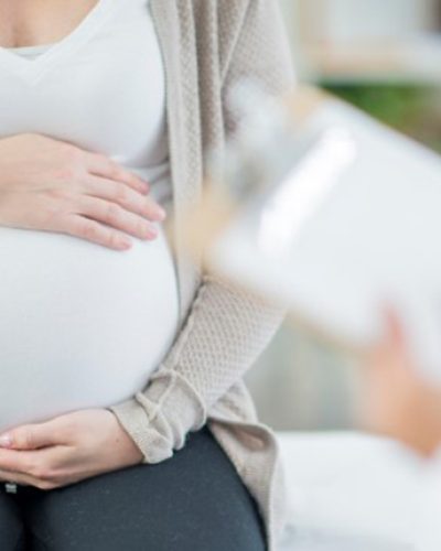 A pregnant Caucasian woman is indoors in a doctor's office. Her female doctor is wearing medical clothing. The woman is holding her stomach which the doctor writes on a clipboard.